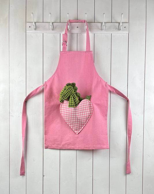 Pink child apron with gingham heart shaped pocket. From Sterck & Co. Dinosaur not included.