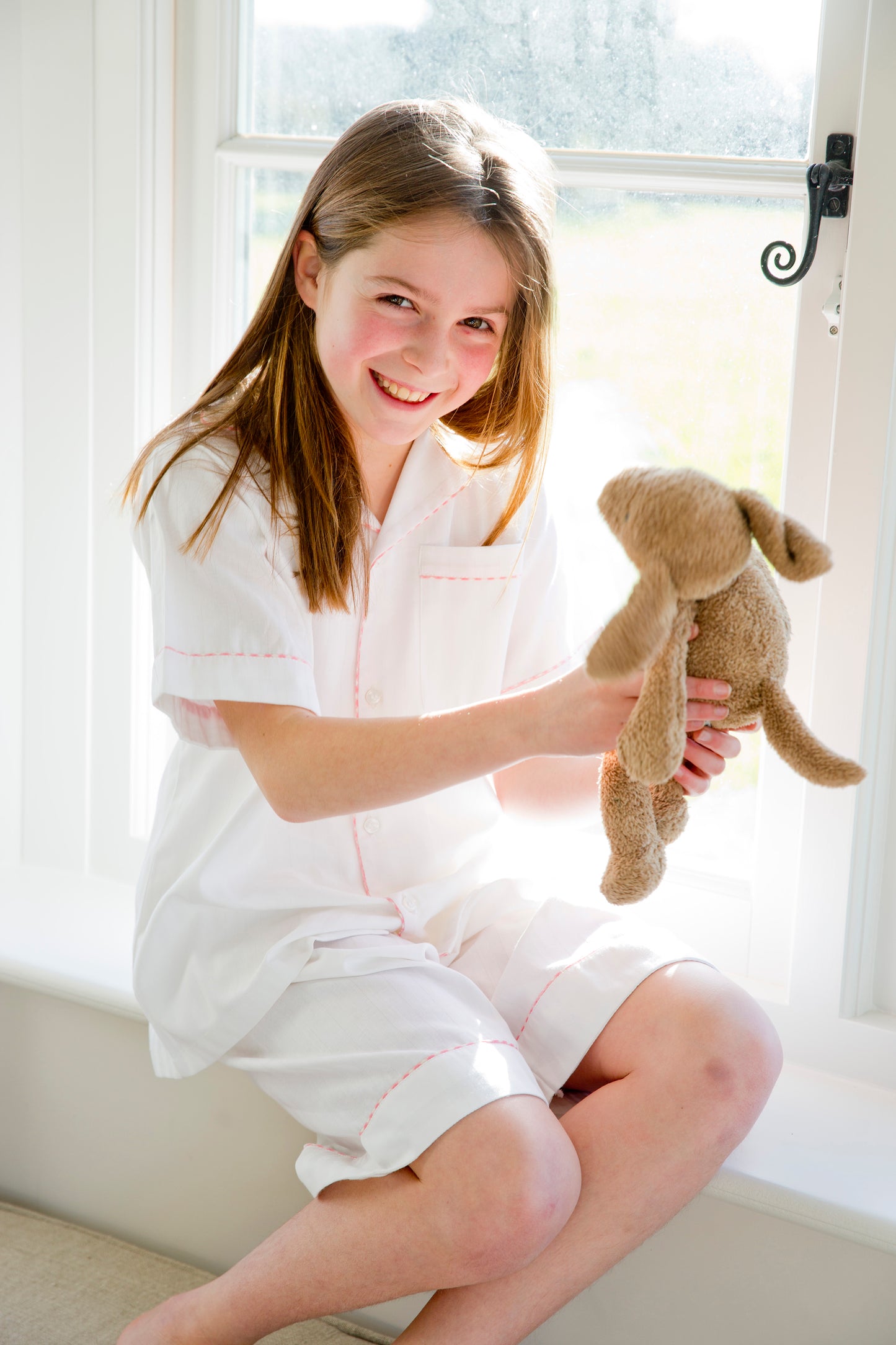 A girl sat on a windowsill with a stuffed toy in classic cotton pyjamas, white with pink piping