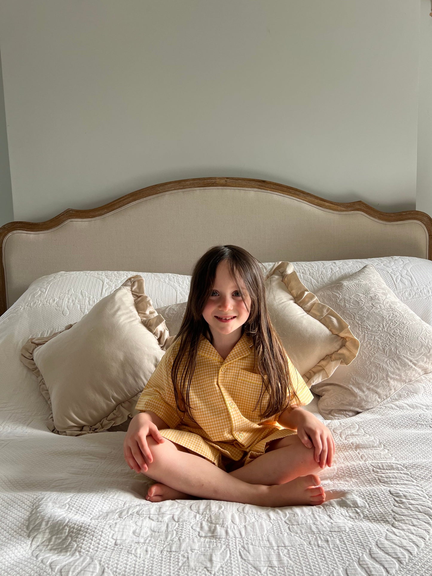 A girl sat on a bed wearing classic Sunny Day yellow gingham cotton pyjamas by TurQuaz.