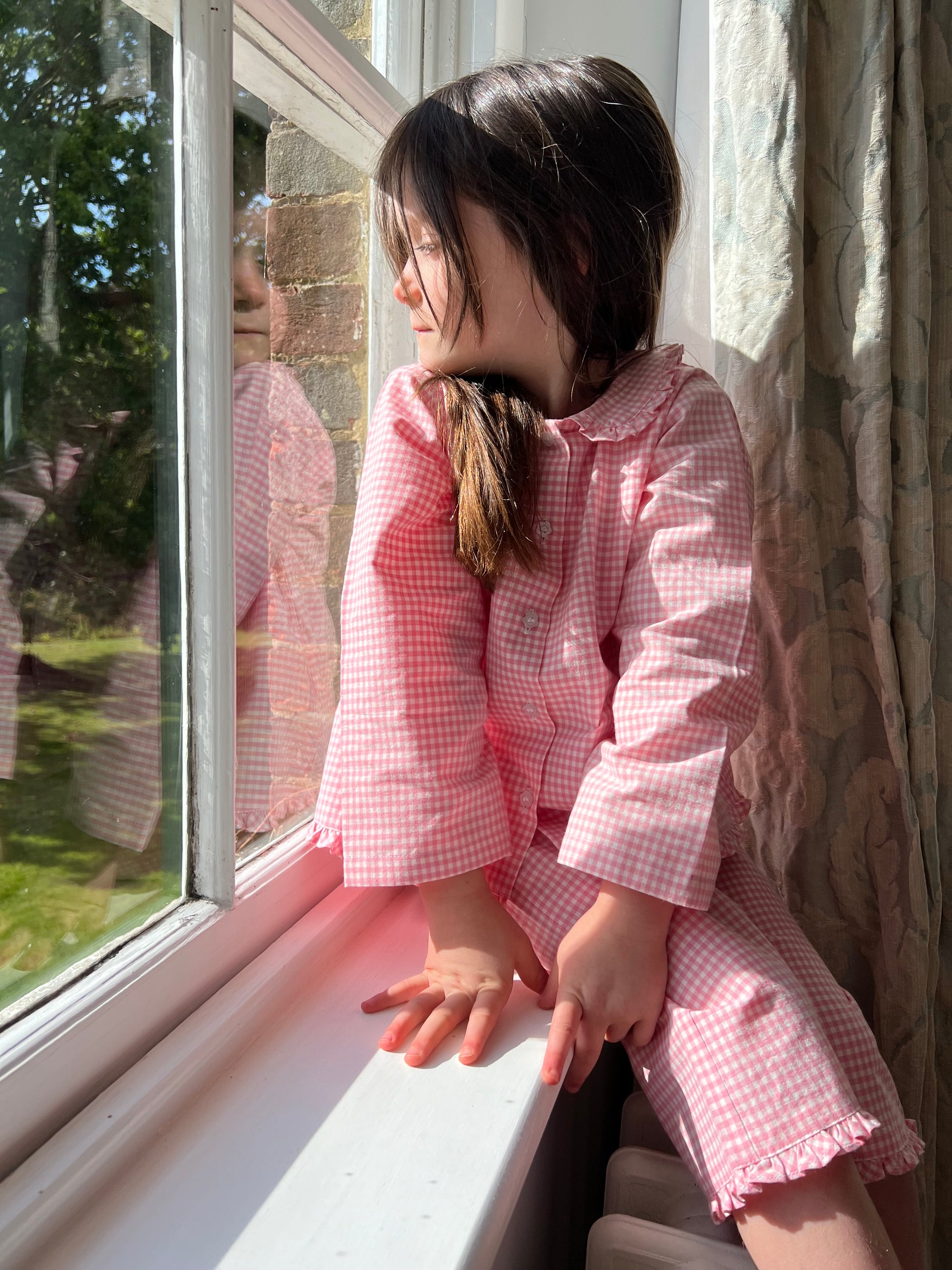 A girl sitting on a windowsill wearing Cotton Candy pink gingham cotton pyjamas by TurQuaz