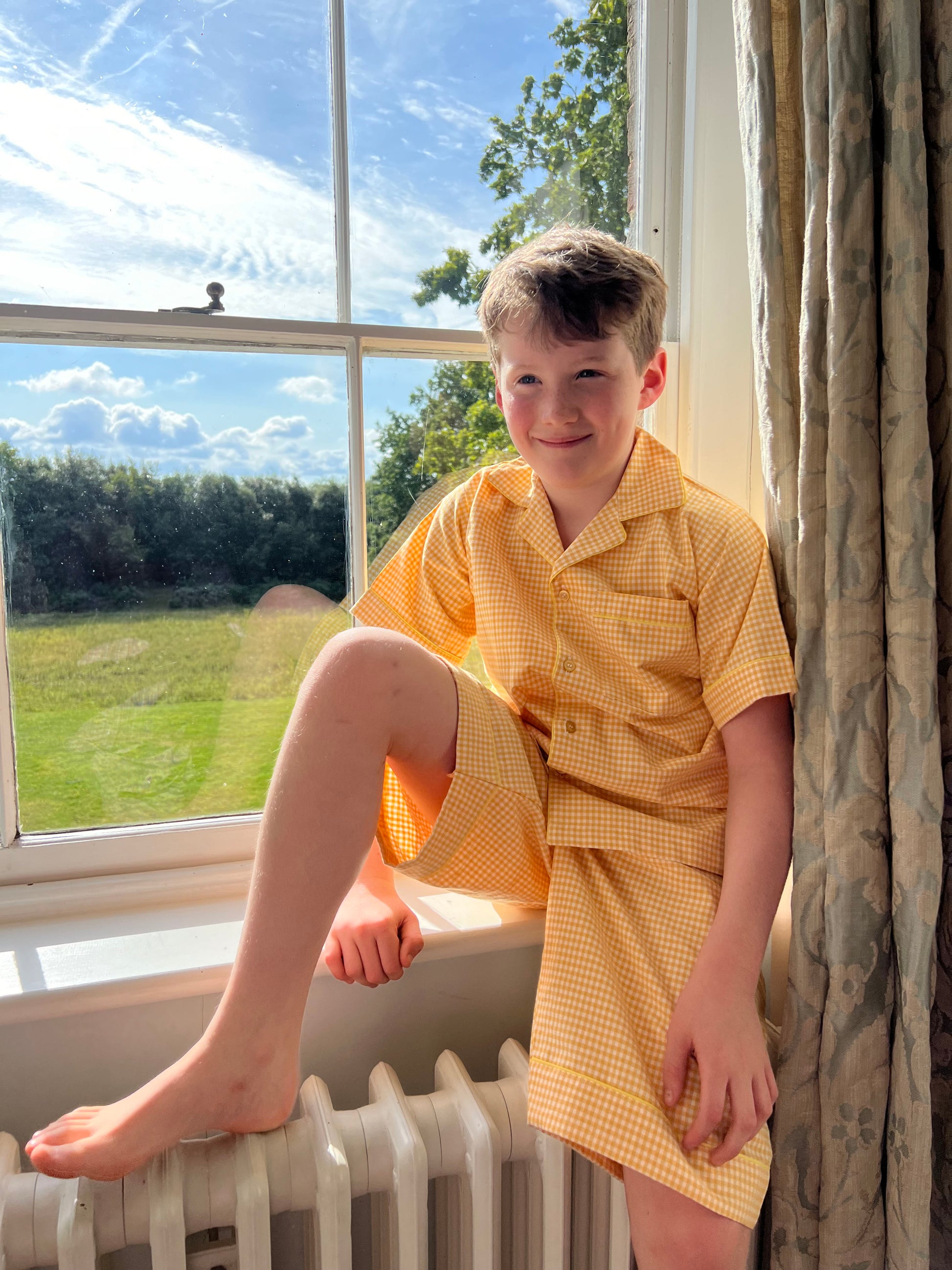 A boy sat on a windowsill wearing classic yellow gingham cotton pyjamas by TurQuaz.