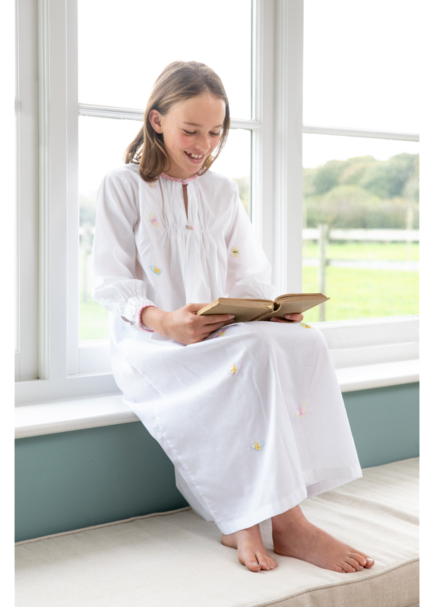 A young girl sits on a window seat in a white nightie, smiling while reading a book. The background shows a grassy field through the window. Designed by TurQuaz the nightie is made from pure cotton fabric with beautiful hand embroidered butterflies on the delicate white fabric.