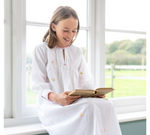 A young girl sits on a window seat in a white nightie, smiling while reading a book. The background shows a grassy field through the window. Designed by TurQuaz the nightie is made from pure cotton fabric with beautiful hand embroidered butterflies on the delicate white fabric.