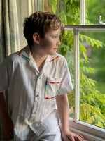 A boy looking out of a window wearing white cotton pyjamas with rainbow detailing on the trims. By TurQuaz.