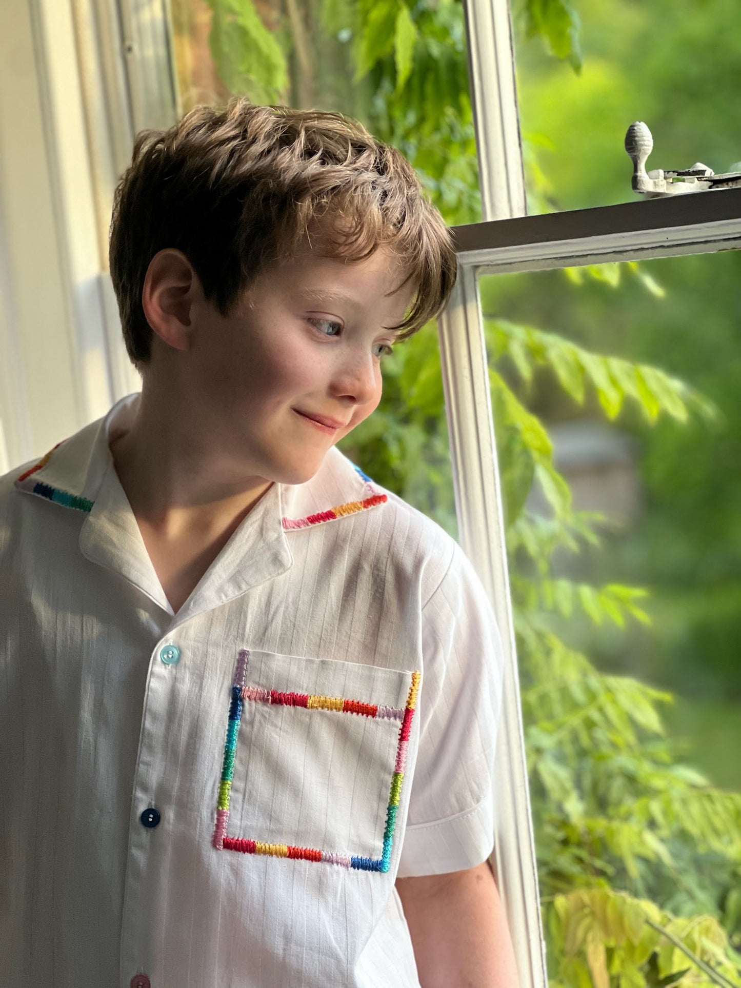 A close up of a boy looking out of a window wearing white cotton pyjamas with rainbow detailing on the trims. By TurQuaz.