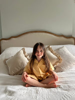 A girl sat on a bed wearing classic Sunny Day yellow gingham cotton pyjamas by TurQuaz.